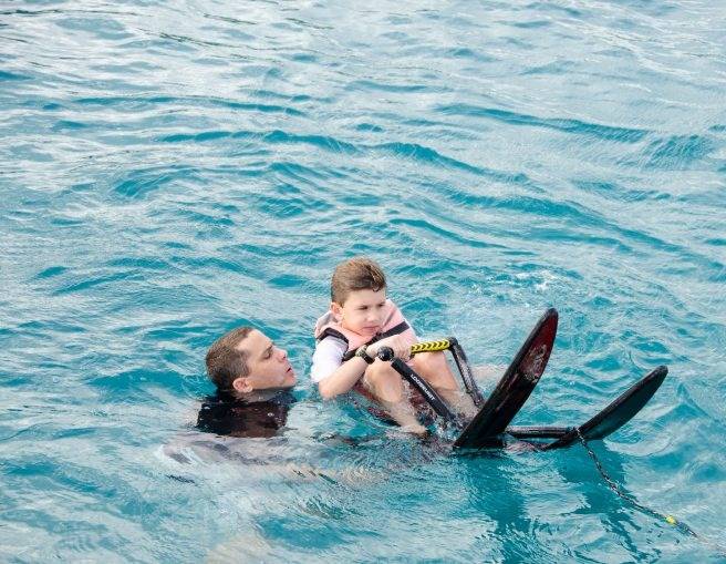 An adult assists a child holding water skis while floating in clear blue water, enjoying the fun and adventure of a luxury yacht charter.