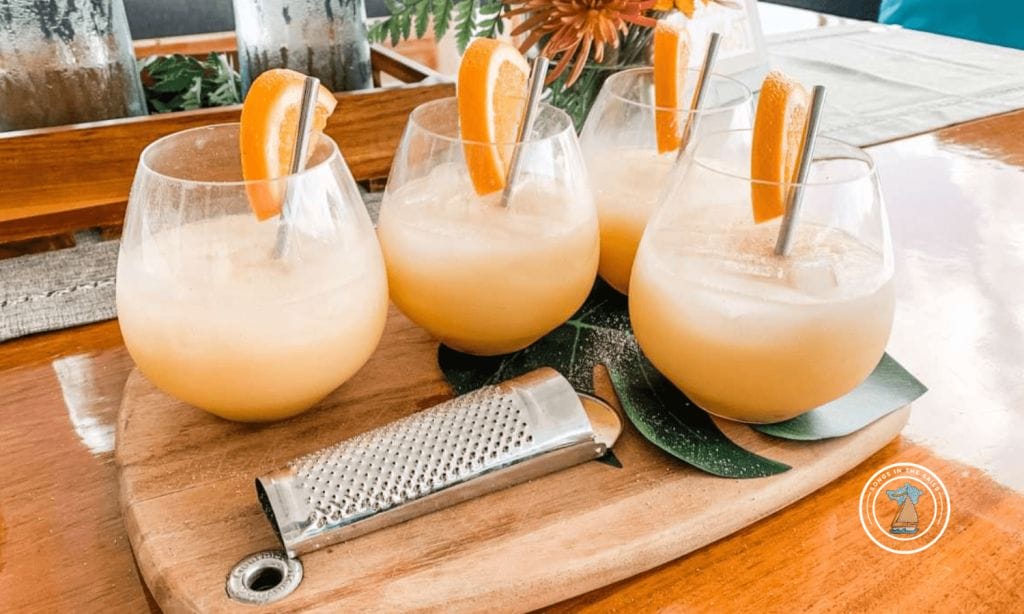 Four glasses of orange-colored drinks with orange slices and metal straws are arranged on a wooden board, next to a metal grater and a decorative leaf.