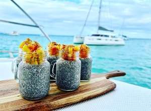 Four jars of chia pudding topped with fruit on a wooden board, set on a crewed yacht charter with the ocean and a sailboat in the background.