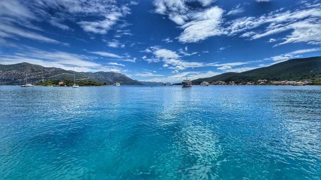 Clear blue water with several boats anchored near a mountainous coastline under a partly cloudy sky.